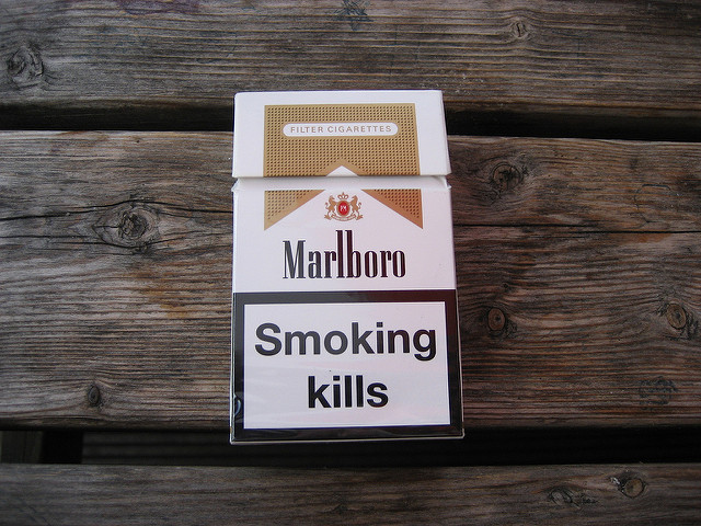 "A pack of Marlboro cigarettes is placed on a wooden surface. The pack has a white and gold design with the Marlboro logo prominently displayed. Below the logo, there is a large warning label that reads 'Smoking kills.'"
