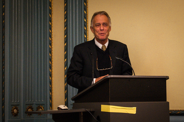 "A person in a suit stands behind a podium equipped with a microphone and some papers. The background features ornate blue and gold paneling."