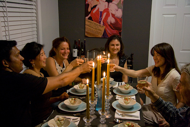 "A group of six people are seated around a dining table, raising their glasses in a toast. The table is set with white plates, bowls of salad, and tall yellow candles in glass holders. Behind them is a painting featuring abstract figures and text. The room has gray walls and white blinds covering a window on the left."