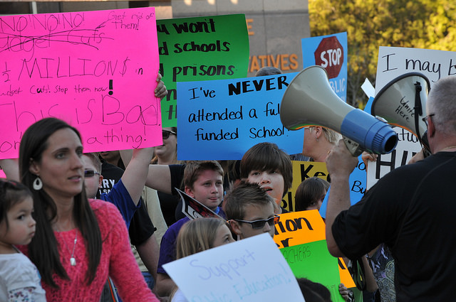 "A group of people are holding various protest signs at a rally. The signs include messages such as 'NO NO NO,' 'Millions $,' 'This is Bad,' 'I've NEVER attended a fully funded school,' and 'Support public education.' One person is speaking into a megaphone."