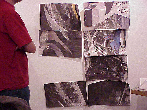 "A person in a red shirt stands facing a white wall with six black-and-white aerial photographs or maps taped to it. The images depict urban areas with buildings and streets. In the top right corner, partial text reads 'LOOKING for an easier READ.'"