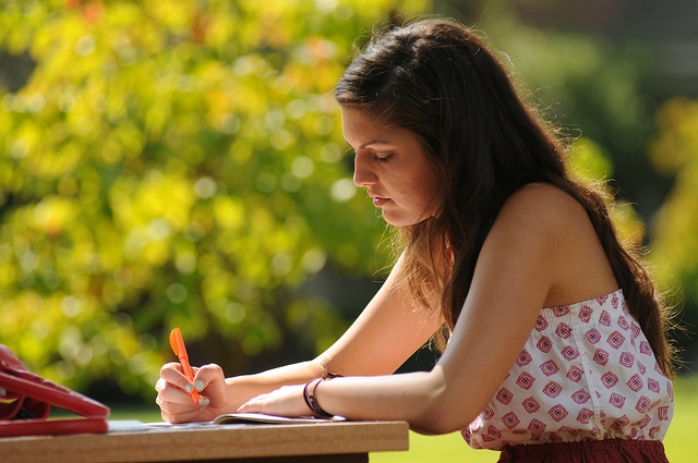 "A person with long hair sits at an outdoor table, writing in a notebook with an orange pen. The background is blurred with green foliage and sunlight. The person is wearing a sleeveless top with a geometric pattern."
