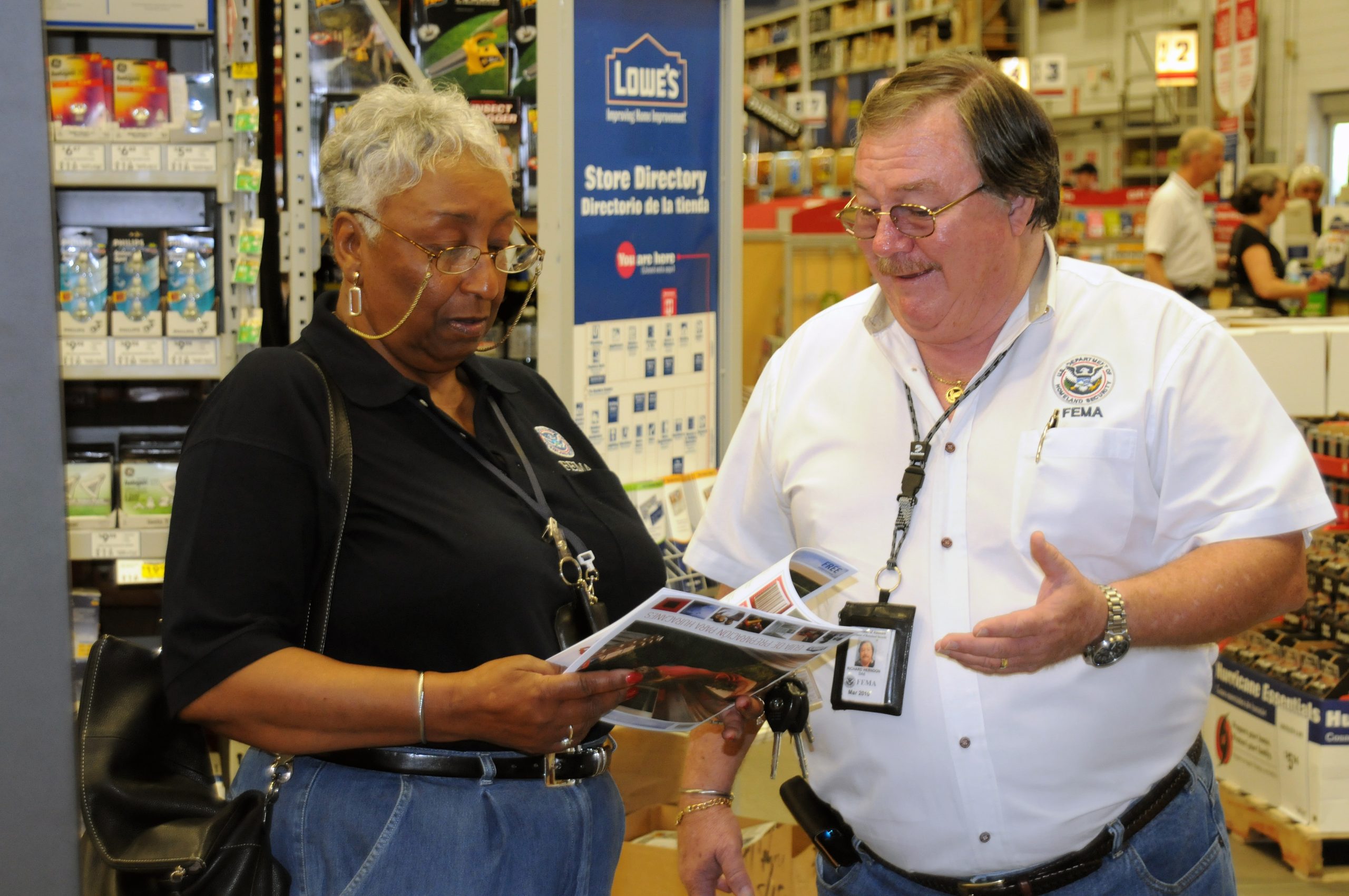 "Two individuals are standing in a store, engaged in conversation. Both are wearing white shirts with logos and name tags. One person is holding a booklet or pamphlet, while the other gestures with one hand. In the background, shelves stocked with various items and a blue sign reading 'Lowe's Store Directory' are visible."