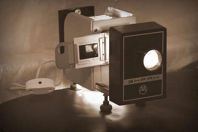 "A vintage film projector labeled 'ЭКРАН' sits on a dark surface with a light shining through its lens. A small white remote control is connected to the projector. The background features a light-colored fabric or curtain."