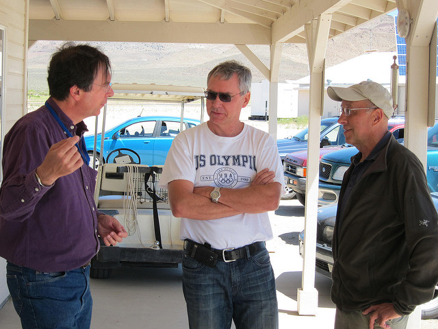 "Three individuals are standing under a covered outdoor area, engaged in conversation. The person on the left wears a purple shirt and blue jeans, gesturing with their left hand. The person in the middle wears a white 'US OLYMPI' t-shirt with arms crossed. The person on the right wears a dark jacket and a beige cap. In the background are several parked vehicles, including a blue car and a golf cart, with mountains visible in the distance."