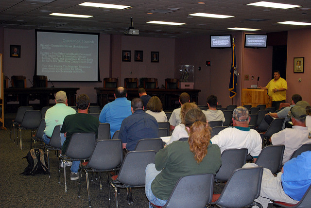 "A group of people are seated in a conference room facing a projector screen displaying a presentation titled 'Operational Hazards.' The slide includes bullet points about hazard identification, assessment, and risk mitigation. Two smaller screens on either side show the same content. The room has rows of gray chairs, and an American flag stands next to a wooden podium where a person is presenting."