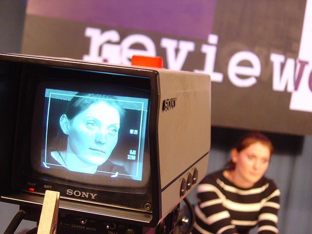 "A close-up of a Sony video camera screen showing a person with their face blurred. In the background, another person with a blurred face is seated in front of a wall with partially visible text reading 'revie w.' The camera screen displays various settings and information."