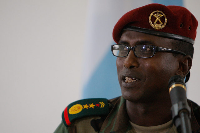 "A person in military uniform speaks into a microphone. The uniform includes a red beret with an insignia and shoulder epaulets featuring stars and other symbols."