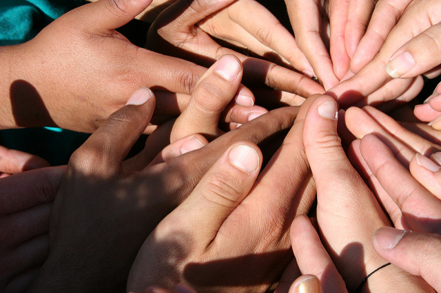 "A close-up image of multiple hands coming together in a circle, with fingers pointing toward the center. The hands, belonging to people of different skin tones, overlap each other, symbolizing unity and teamwork."