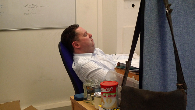 "A person sits in an office chair, leaning back and facing away from the camera. A whiteboard with partially legible text is mounted on the wall. On the desk in front of the person are various items, including a can of SlimFast, a glass jar, and other small objects. A blue curtain or divider partially obscures the right side of the image."