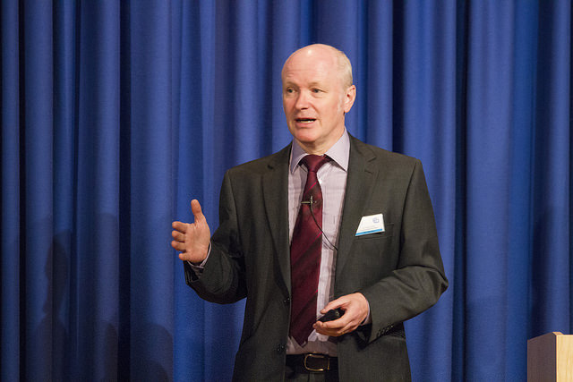 "A person in a suit and tie stands in front of a blue curtain, giving a presentation. They hold an object in their right hand and gesture with their left. A microphone is clipped to their shirt, and a name tag is visible on the left side of their suit jacket."