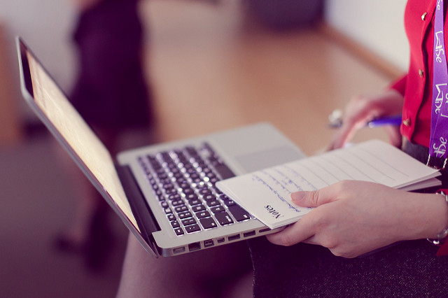 "A person sits with a laptop on their lap and a notepad in their left hand. They are typing or working on the laptop while holding a pen in their right hand. The notepad contains handwritten notes. In the background, a blurred figure and part of a room with wooden flooring are visible."