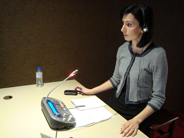 "A person stands at a podium equipped with a microphone and headset, holding a remote control. On the table are a water bottle, some papers, and an electronic device with buttons and a display screen. The background is plain and dark."