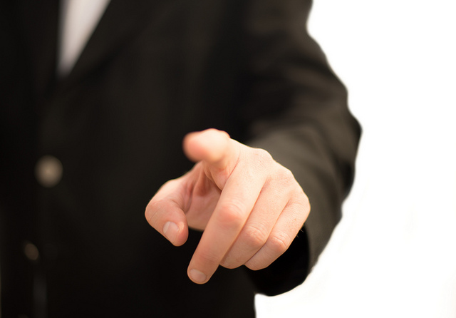 "A person in a black suit and white shirt points directly at the camera with their index finger extended. The background is plain white, emphasizing the hand and suit."