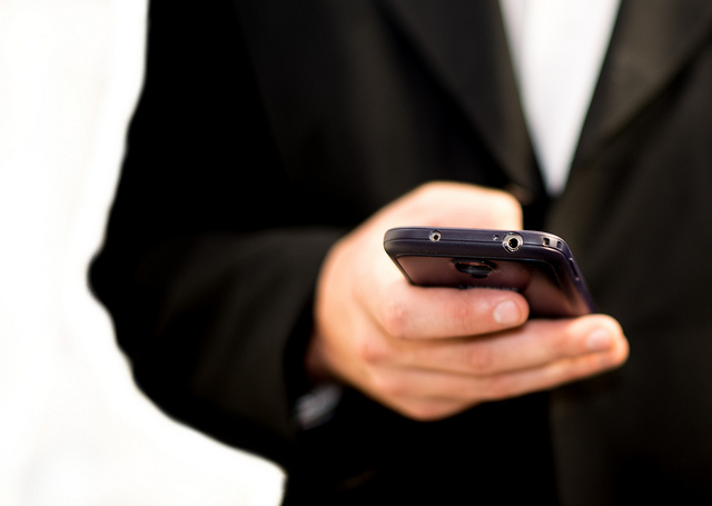 "A person in a black suit holds a smartphone in one hand. The focus is on the hand and the phone, which has a visible headphone jack and camera lens on its top edge. The background is blurred."