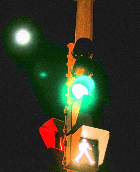 "A traffic light at night displays a green light and a pedestrian signal with an illuminated walking figure. The background is dark, with two bright circular lights visible in the upper left corner."