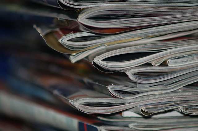 "Close-up of a stack of magazines or newspapers with slightly curled and overlapping pages, emphasizing the texture and layers of the paper."