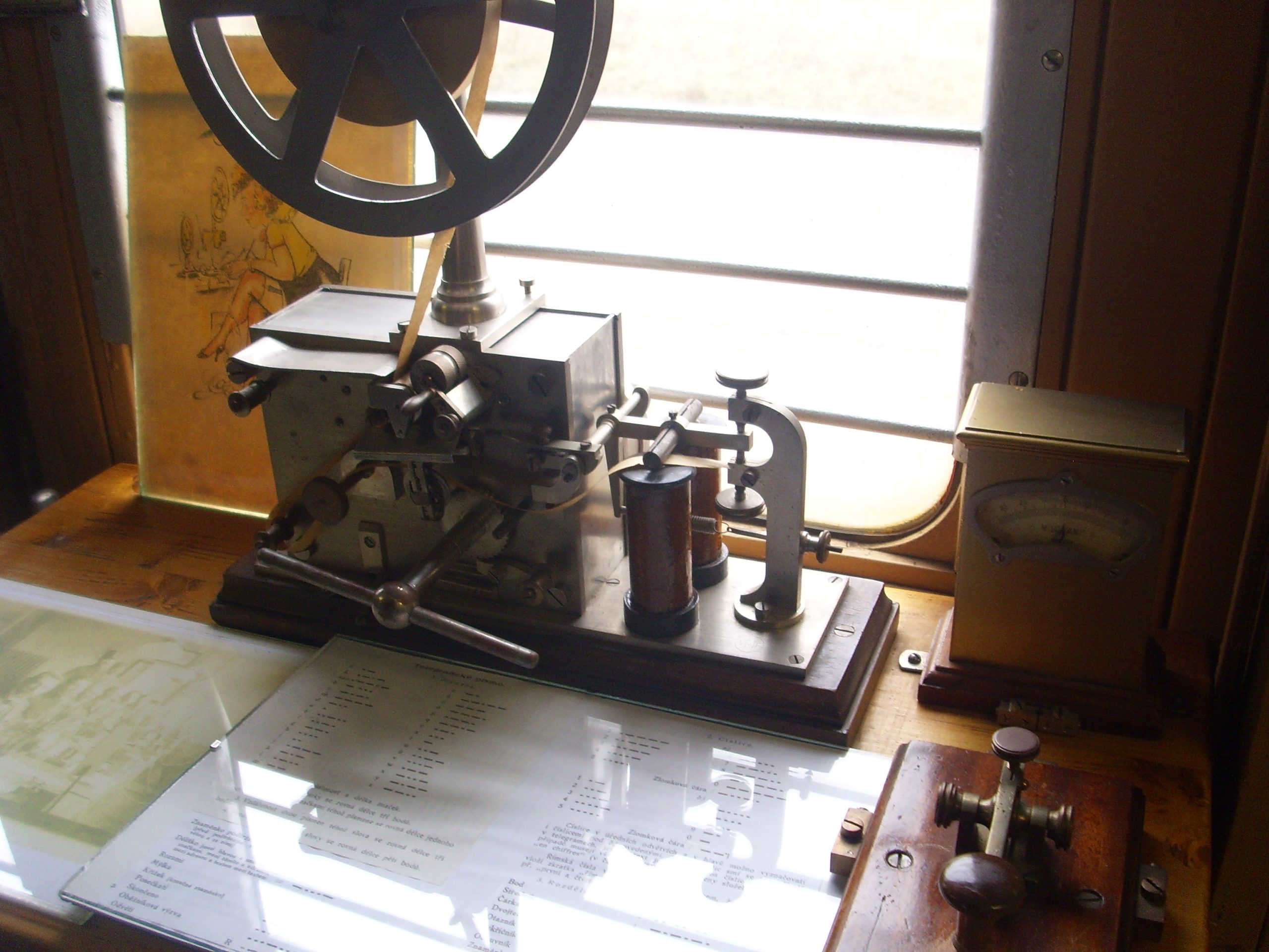 "An old telegraph machine with mechanical components including a large wheel, levers, a cylindrical part, and a gauge on the right. It is placed on a wooden surface with a glass-covered document beneath it. A window with horizontal bars and an illustration are visible in the background."