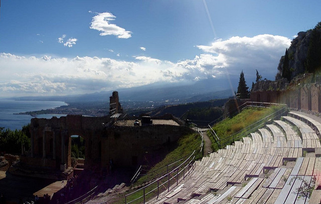 "An ancient amphitheater with semi-circular stone seating overlooks a scenic coastline and sea, with mountains in the background under a partly cloudy sky. The ruins of the stage area are visible on the left, and greenery surrounds parts of the structure."