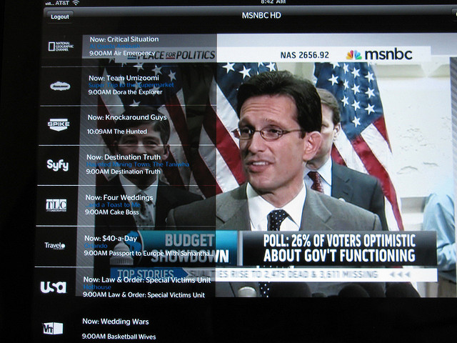 "A television screen displays a news broadcast on MSNBC HD, showing a man speaking at a podium with American flags in the background. His face is blurred. A news ticker reads, 'POLL: 26% OF VOTERS OPTIMISTIC ABOUT GOV'T FUNCTIONING.' On the left, a TV guide lists programs on channels like AMC, Spike, Syfy, TLC, USA Network, and WE tv, including 'Critical Situation,' 'Knockaround Guys,' 'Despicable Me,' 'Four Weddings,' and 'Basketball Wives.'"