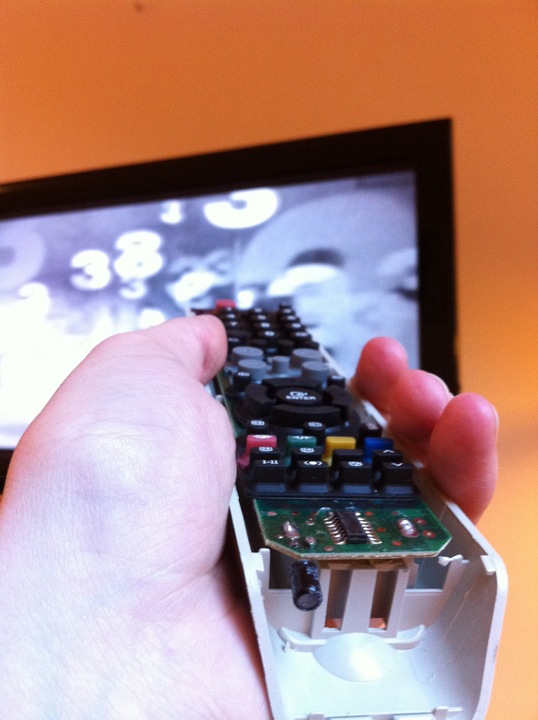 "A person holds a disassembled remote control pointed at a television screen. The remote's casing is partially removed, revealing the circuit board and internal components. The TV screen in the background shows a black and white image with various numbers and shapes."