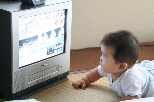 "A baby lies on a tatami mat floor, watching a small Sony television placed on the floor. The screen shows a colorful image, possibly from an animated show or video game. The baby is wearing a white shirt and has short dark hair."