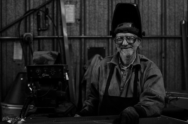 "A black and white image of a person wearing welding gear, including a lifted welding helmet. Their face is obscured by a blurred rectangle. They stand in front of welding equipment inside an industrial workshop with corrugated metal walls, with various tools and machinery in the background."