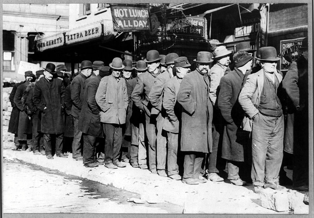 A black and white historical photograph showing a long line of men standing on a sidewalk, waiting in front of businesses. The men are dressed in heavy coats and hats, suggesting cold weather. Signs above the businesses read "HOT LUNCH ALL DAY" and "FREE GRANT'S EXTRA BEER." The image captures a scene from the Great Depression era, highlighting the economic hardship as people wait for food or assistance.