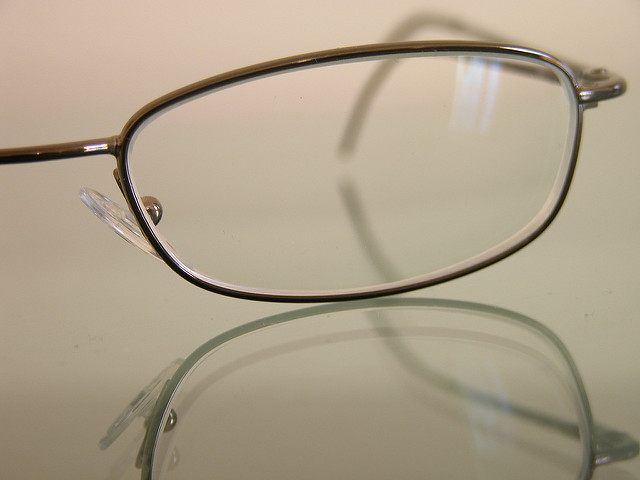 A close-up image of a pair of eyeglasses with a thin metallic frame. The glasses are placed on a reflective surface, creating a clear reflection of the frame below. The background is blurred and neutral in color, highlighting the eyeglasses as the main subject.