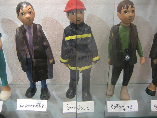 The image shows three figurines displayed in a glass case. Each figurine represents a different profession and has a blurred face. From left to right, the professions are labeled as "inşaatçı" (construction worker), "bomber" (firefighter), and "fotograf" (photographer). The construction worker is dressed in a suit with a briefcase, the firefighter is wearing a fire-resistant suit with reflective stripes and a red helmet, and the photographer is holding a camera around the neck.