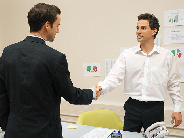Two individuals are shaking hands in an office setting. Both individuals have their faces blurred for privacy. One person is wearing a dark suit, while the other is dressed in a white shirt and black pants. The background features various charts and graphs pinned to the wall, indicating a business or professional environment. A desk with papers, a phone, and a yellow chair is also visible in the foreground.