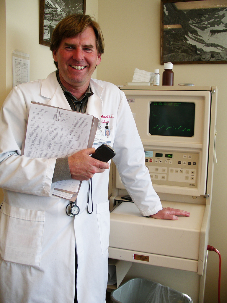 A medical professional wearing a white lab coat stands in front of medical equipment, holding a clipboard and a smartphone. The person's face is obscured. The equipment behind the professional includes a monitor displaying green waveform lines, likely representing heart activity. There are framed pictures on the wall and various items on top of the equipment, including a brown bottle.