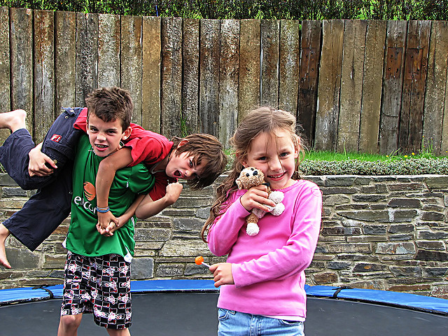 Three children are playing on a trampoline in a backyard. One child is wearing a green shirt and holding another child, who is wearing a red shirt, over the shoulder. The third child, wearing a pink sweater, stands in front holding a small stuffed animal and an orange lollipop. The background features a wooden fence and stone wall.