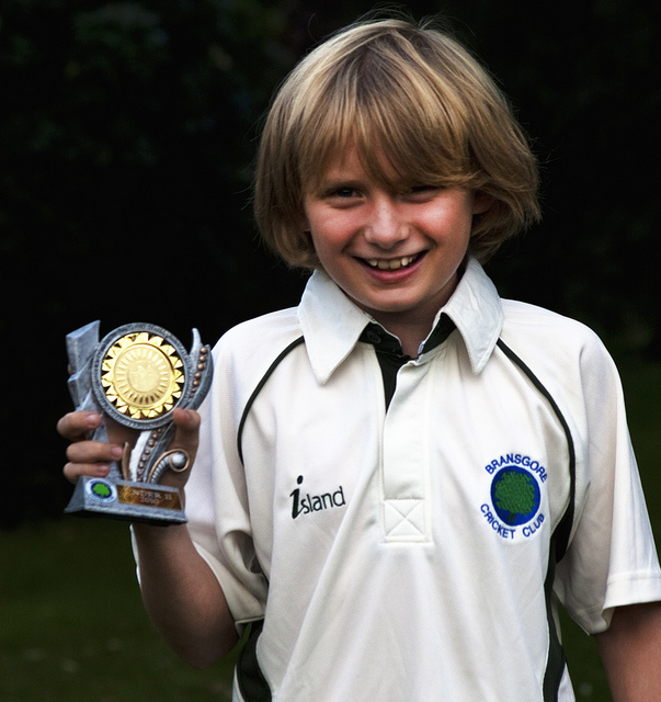 "A young person with blond hair, wearing a white cricket shirt with the logo 'BRANSGORE CRICKET CLUB' and holding a trophy in the left hand. The face is blurred out for privacy. The background is dark and out of focus, highlighting the person and the trophy."