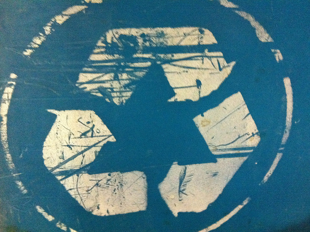 "A blue surface with a white recycling symbol that appears worn and scratched. The recycling symbol consists of three arrows forming a triangle, indicating the concept of recycling and environmental conservation. The scratches and wear on the surface suggest it has been used frequently or exposed to rough conditions."