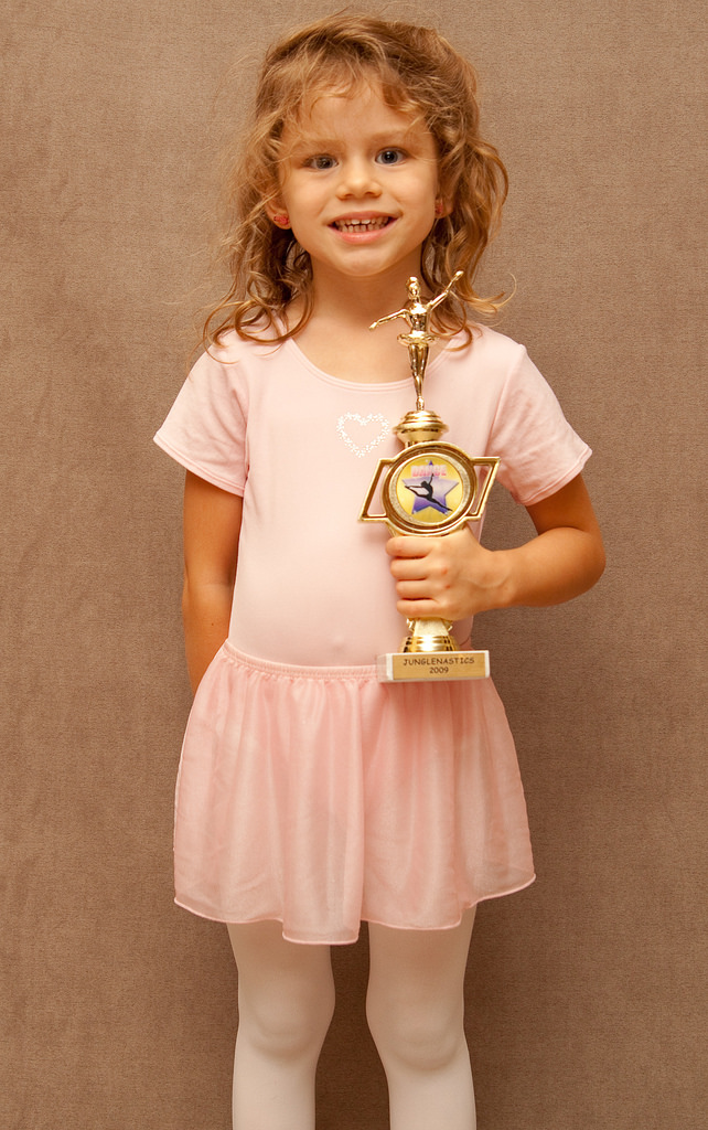 "A young child with long, curly hair stands against a plain background, holding a gold trophy with both hands. The child is wearing a light pink dress with short sleeves and white tights. The child's face is pixelated for privacy."
