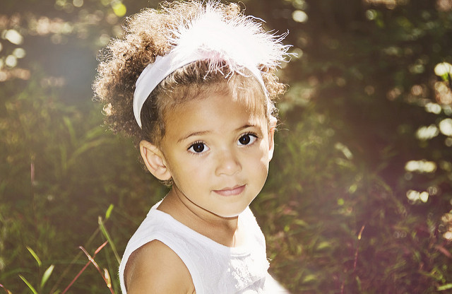 "A young child with curly hair wearing a white headband with a feather and a white sleeveless dress is standing outdoors in a sunlit area with green grass and foliage. The child's face is blurred for privacy. The sunlight creates a soft, warm glow around the child, highlighting the natural setting."