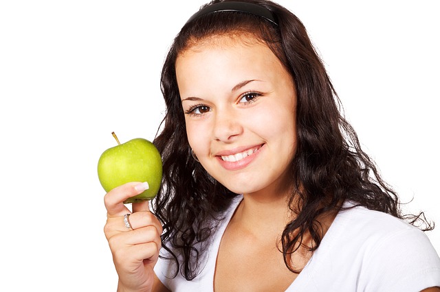 "A person with long, curly hair is holding a green apple in the right hand. The person's face is blurred out, and they are wearing a white shirt. The background is plain white."
