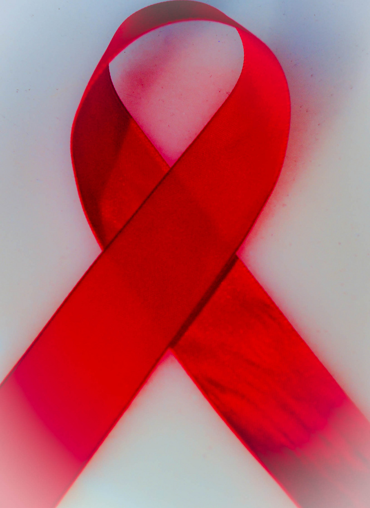 "A red ribbon folded into a loop, symbolizing awareness and support for various causes such as HIV/AIDS awareness. The ribbon is set against a light background with soft shadows and gradients, giving it a three-dimensional appearance."