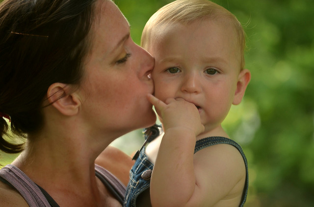 "An adult holding a baby outdoors. The adult is wearing a sleeveless top, and the baby is dressed in denim overalls. Both faces are blurred out. The background is green, suggesting they are in a natural setting like a park or garden."