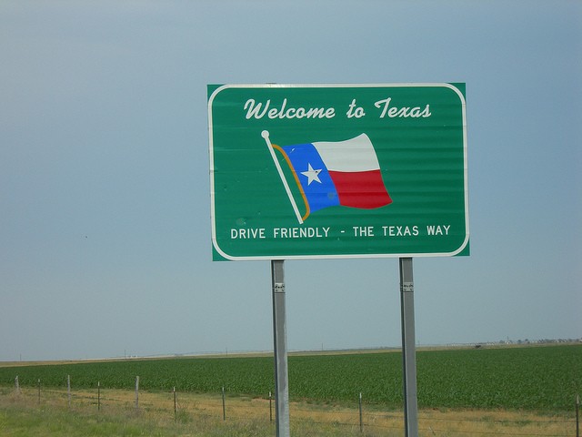 "A green road sign with white text and a depiction of the Texas state flag. The sign reads 'Welcome to Texas' at the top, followed by an image of the Texas flag, and below it says 'Drive Friendly - The Texas Way.' The sign is mounted on two metal poles and is situated in front of a vast field with a clear blue sky in the background."