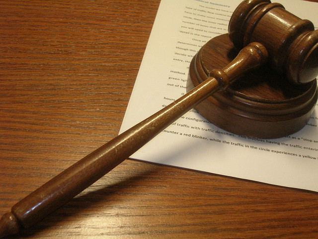 "A wooden gavel rests on a sound block on a wooden surface. Underneath the gavel, there is a piece of paper with printed text, partially visible but not fully readable. The image suggests a legal or judicial context."