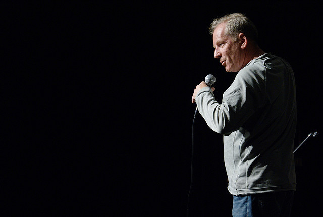 "A person with a blurred face is standing on a dark stage, holding a microphone in the right hand. The person is wearing a light gray long-sleeve shirt and blue jeans. The background is completely black, suggesting that the stage is dimly lit or the photo was taken with a spotlight focused on the person."