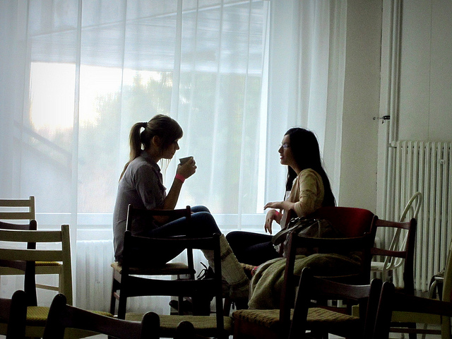"Two people are sitting on chairs in a room with large windows covered by sheer white curtains. They appear to be engaged in conversation, with one person holding a cup. The room has several empty chairs and a radiator is visible against the wall."