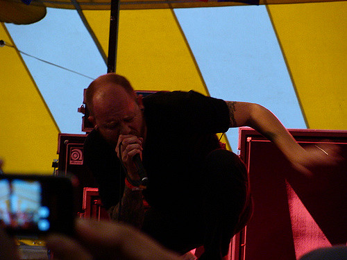 "A person is performing on stage under a yellow and blue striped tent. The person's face is obscured by a pixelated block. The person is wearing a black outfit and has tattoos on the left arm. There are red amplifiers or speakers behind the person, and an audience member's hand holding a smartphone can be seen in the foreground, capturing the performance."