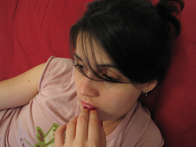 "A person with dark hair is lying on a red surface, wearing a light pink shirt with a green design. The person's face is blurred out, and the hand is near the chin. The person has an earring in the left ear."