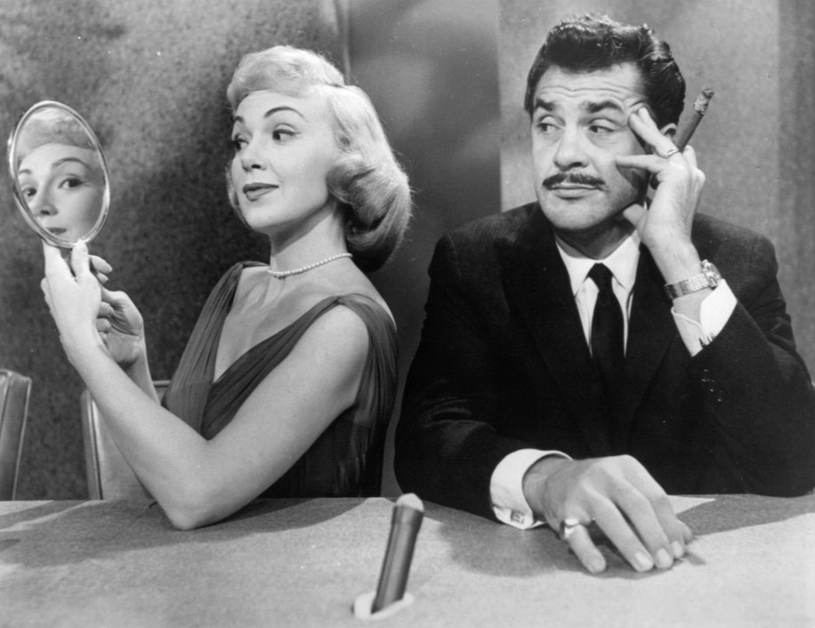 "A black and white photograph shows two individuals sitting at a table. The person on the left is holding a small round mirror in one hand and appears to be adjusting their hair with the other hand. The person on the right is wearing a suit and tie, holding a cigar in one hand, and has their other hand resting on the table. A microphone is placed on the table in front of them. Both individuals' faces are obscured by gray rectangles."
