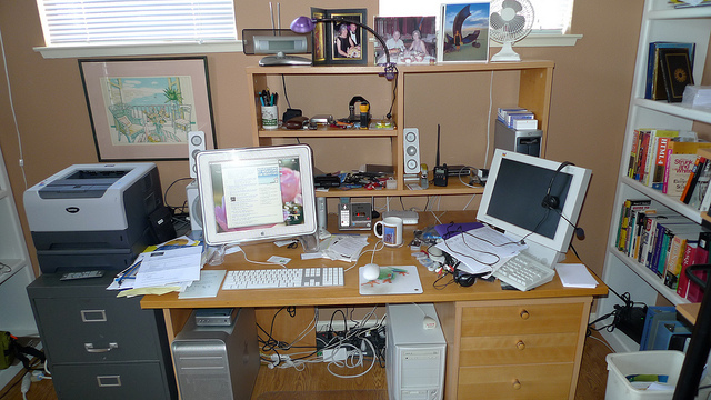 "A cluttered home office desk with two computer monitors, a printer, various office supplies, and bookshelves filled with books. The desk has papers, a keyboard, a mouse, and other electronic devices scattered across it. There are framed pictures on the wall above the desk and a fan on the top shelf."