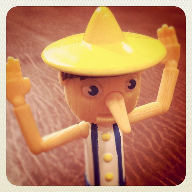 "A close-up photograph of a small plastic toy figure resembling Pinocchio. The figure has a long nose, wide eyes, and is wearing a yellow hat and blue suspenders over a white shirt with yellow buttons. The toy's arms are raised upwards. The background is blurred, showing a wooden surface."