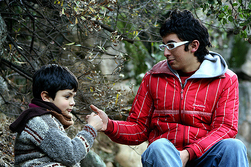 "An adult and a child are sitting outdoors in a natural setting with trees and bushes around them. The adult, wearing a red jacket, is seated on the right side of the image, while the child, wearing a gray sweater with patterns, is seated on the left. They appear to be engaged in an interaction where they are holding hands or passing something between them."
